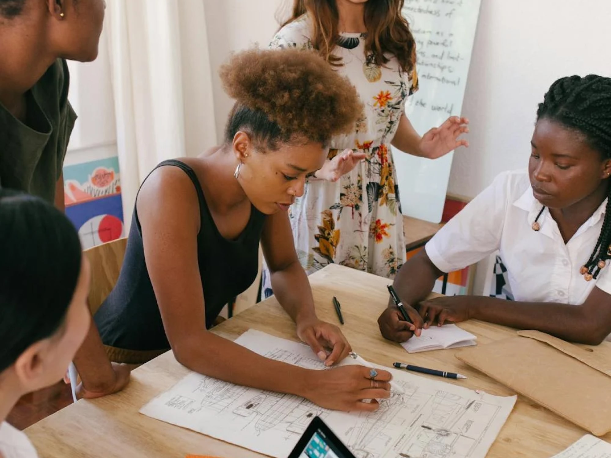 Four people sit and one stands around a table discussing papers and diagrams, with pens, notebooks, and a whiteboard in the background.