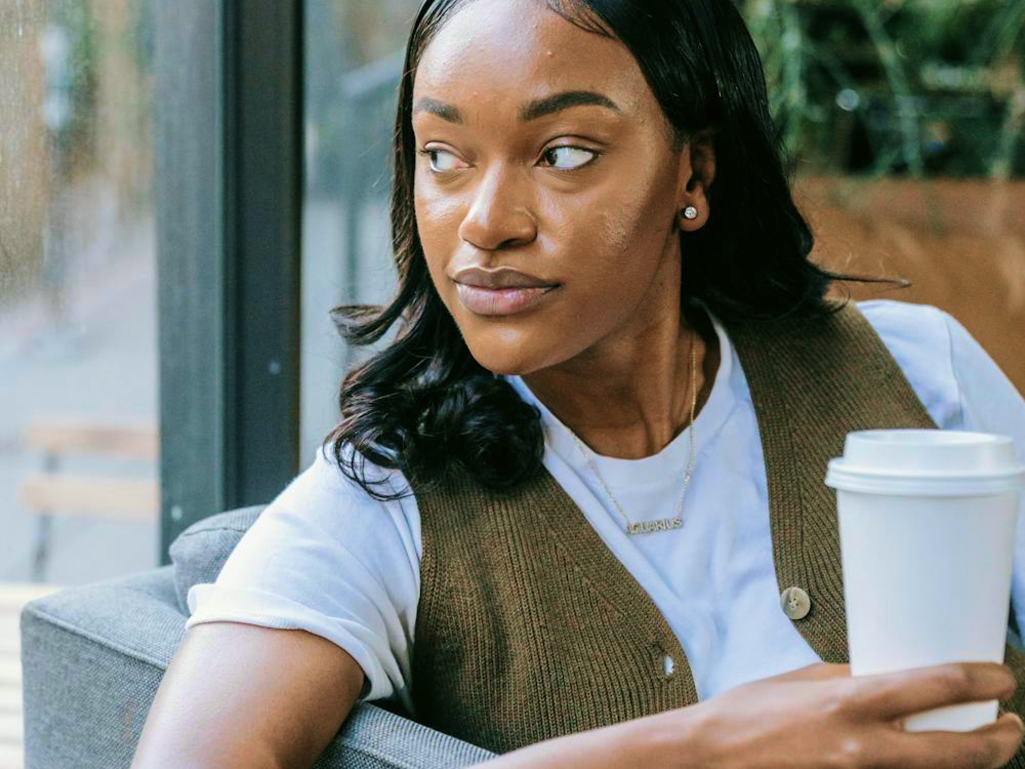A woman with long dark hair sits indoors, holding a takeaway coffee cup and looking to her right. She wears a white shirt, a brown vest, and a necklace.