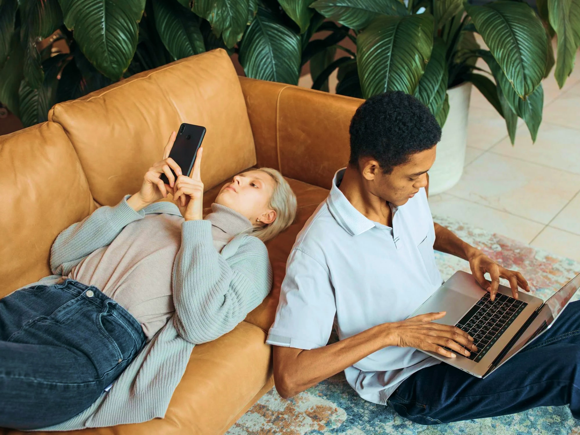 A woman lies on a couch using her phone while a man sits on the floor beside her working on a laptop. Large green plants are in the background.